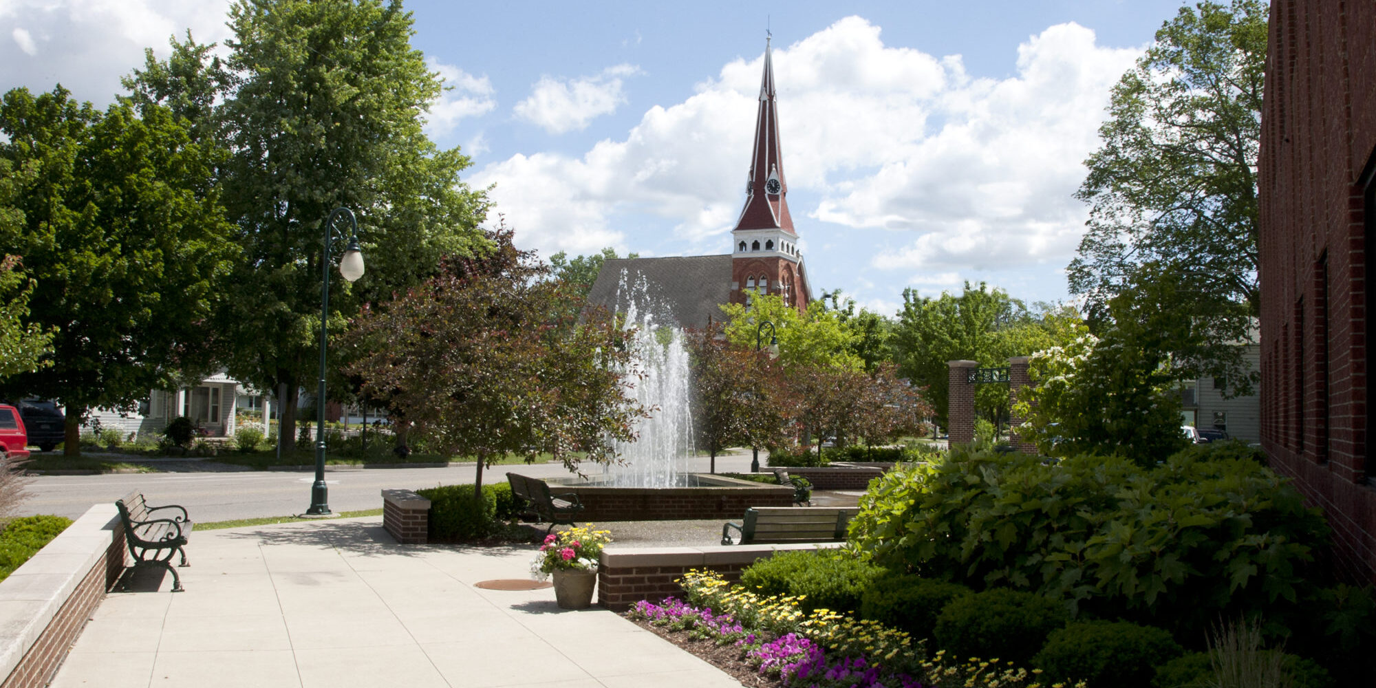 Sunny plaza with fountain, flowers, and church spire.